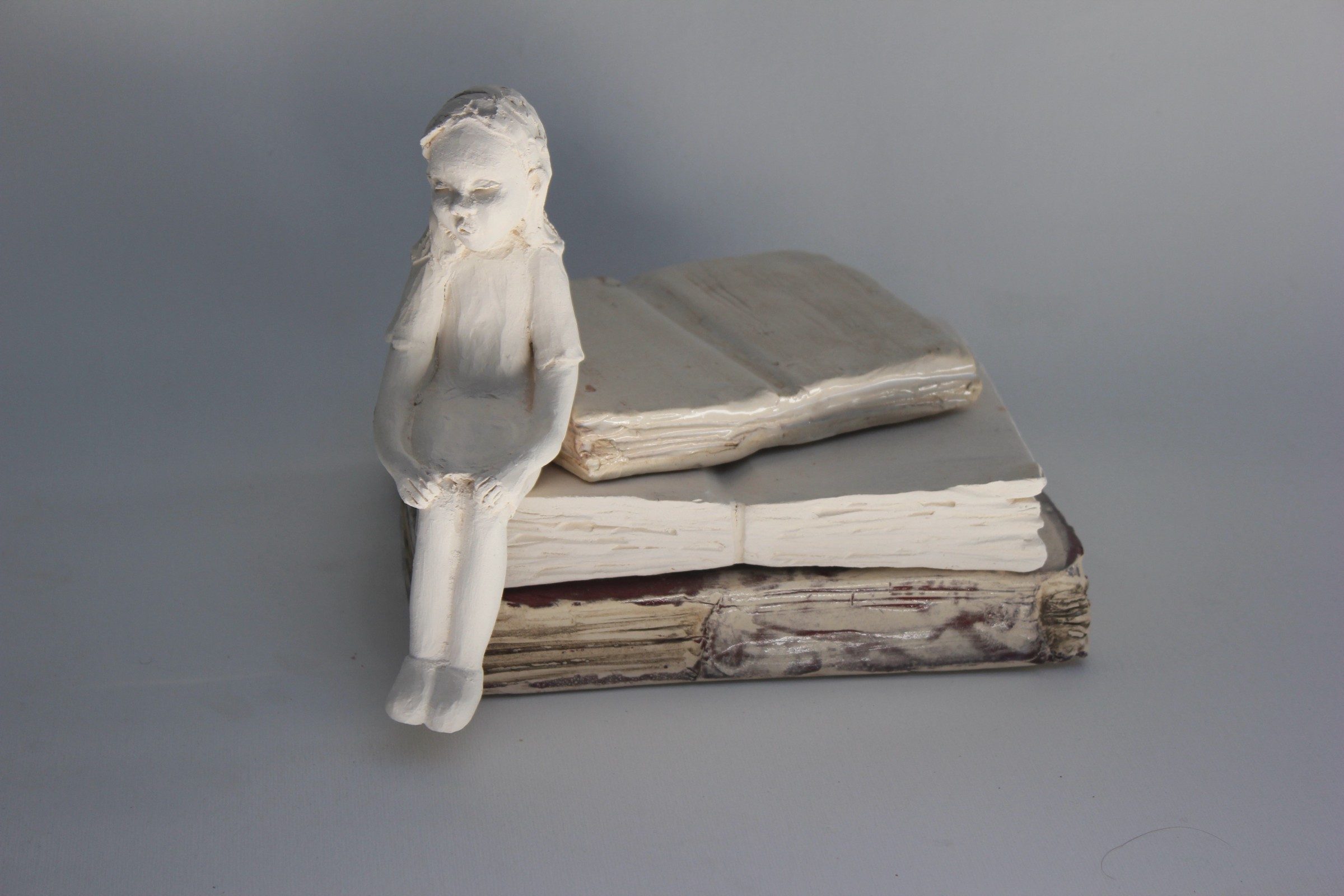 Escultura cerámica de una niña sentada sobre un libro, serie Identità